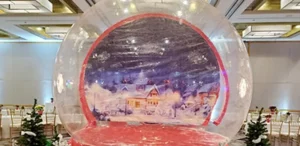 Giant inflatable snow globe with a snowy village backdrop set up in a ballroom between small Christmas trees