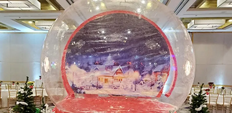 Giant inflatable snow globe with a snowy village backdrop set up in a ballroom between small Christmas trees