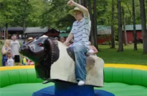 Young rider in a striped shirt holding on with one arm up on a brown and white mechanical bull inside a padded green and yellow inflatable ring at an outdoor picnic