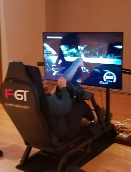 Guest seated in a racing simulator seat with a steering wheel, driving a race on a large mounted screen