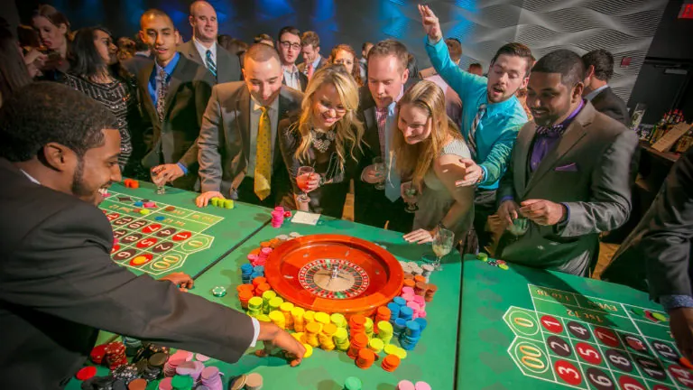 Guests in suits and dresses crowding around a roulette wheel and layout covered in colorful chips