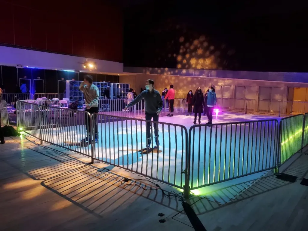 Skaters on a synthetic ice rink at night with colored lighting and metal barricades