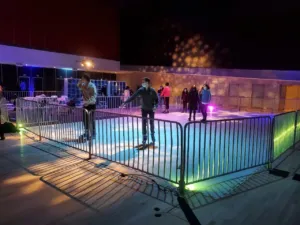 Skaters on a synthetic ice rink at night with colored lighting and metal barricades