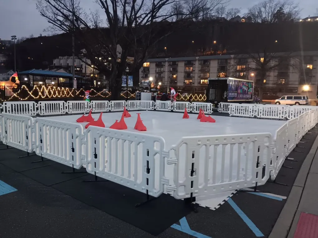Portable synthetic ice skating rink with white barrier fencing set up in a parking lot at dusk